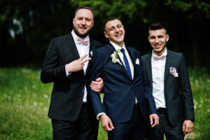 Imagem de padrinhos de casamento ao lado do noivo, todos usando ternos pretos elegantes, com expressões de felicidade e confraternização.